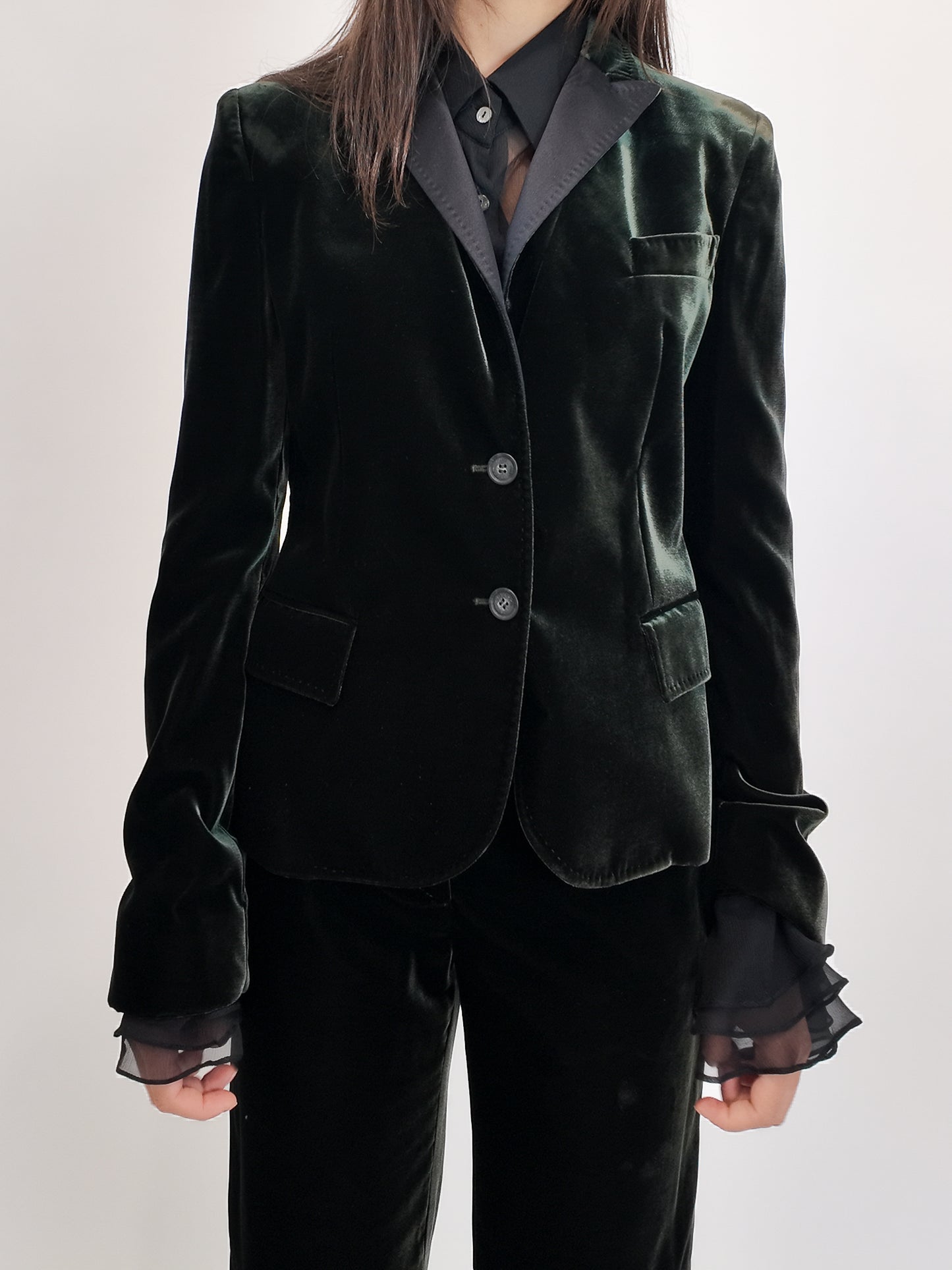 Green velvet blazer with sheer ruffled blouse underneath on a plain background