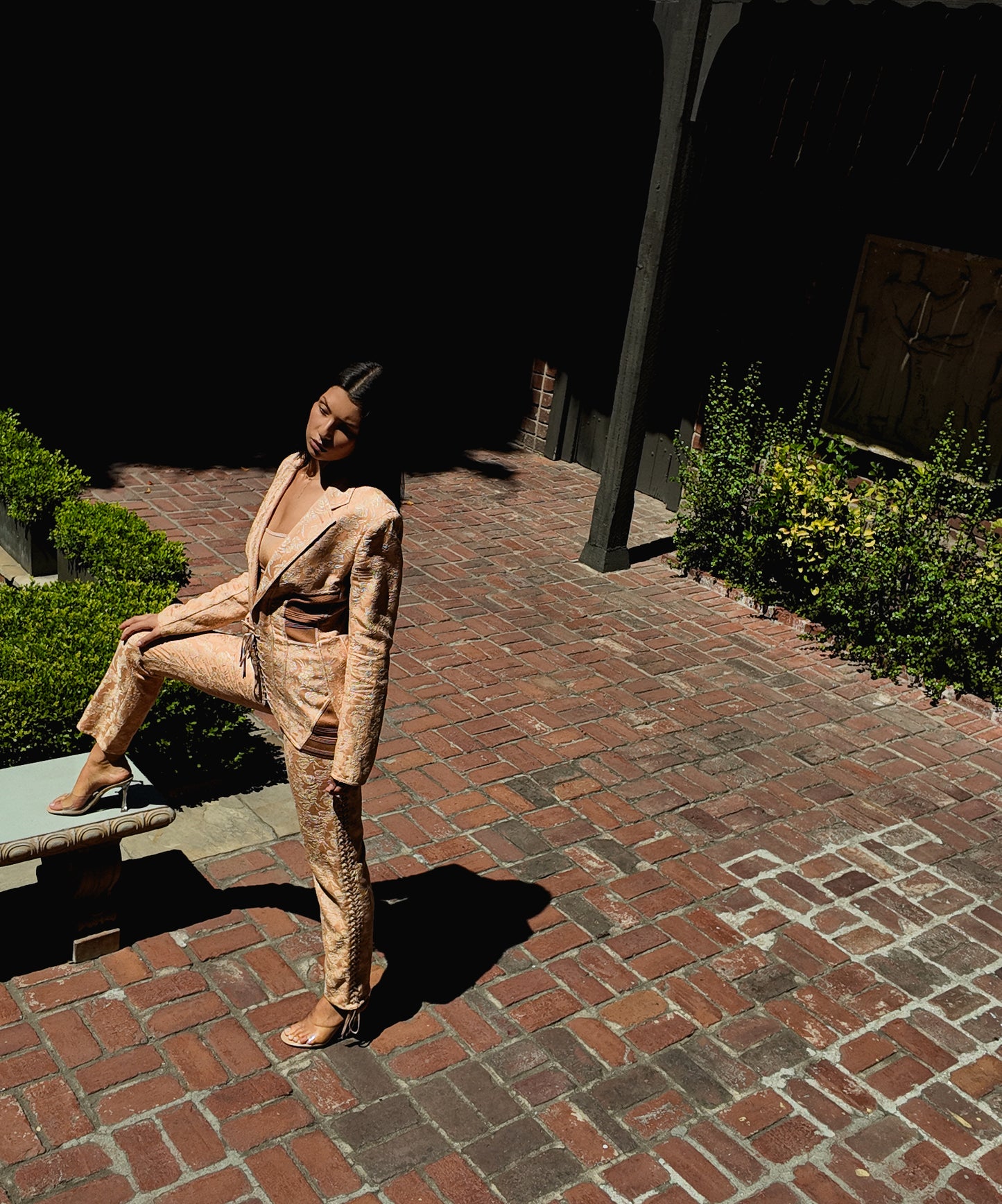 Person in a nude corseted suit sitting on a stone bench in a garden setting