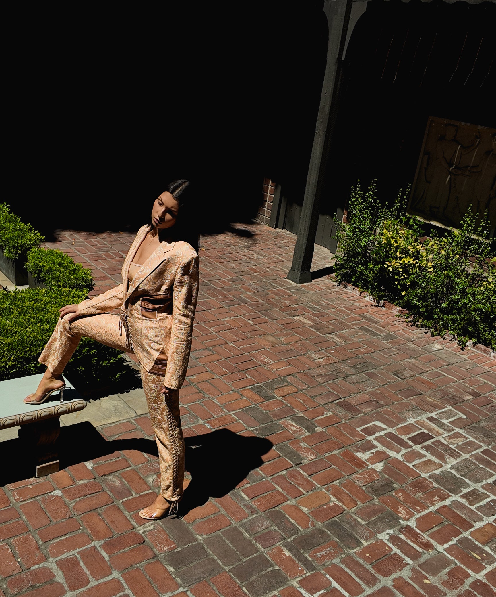 Person in a nude corseted suit sitting on a stone bench in a garden setting
