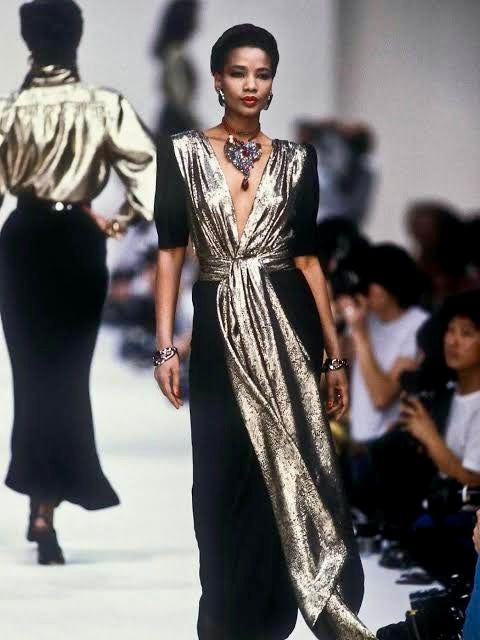 Model wearing a gold and black dress on a runway with blurred audience in the background