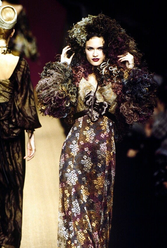 A christian Lacroix 1995 runway photo of a model wearing a voluminous, dark floral-patterned gown with heavily textured, fluffy dark sleeves and an oversized dark feathered or fur-like headpiece, walking on a fashion show.
