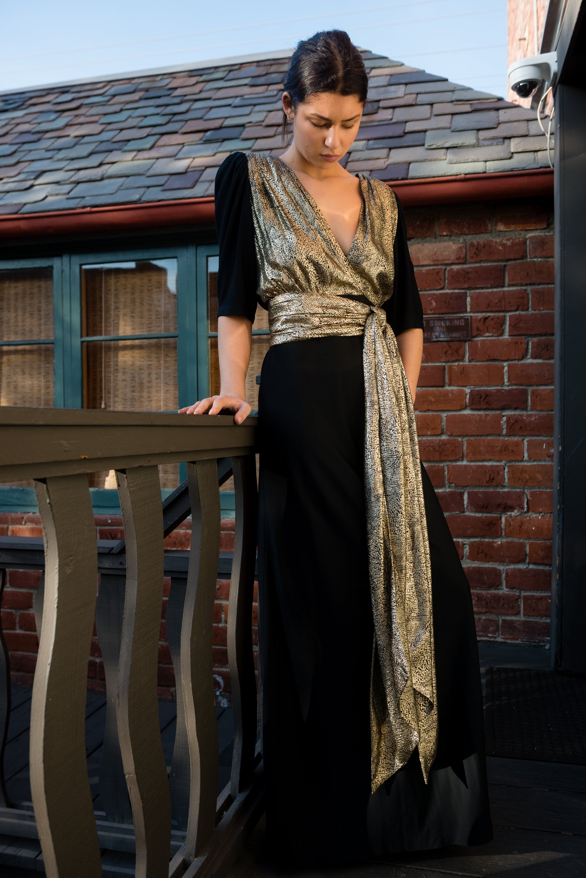Woman in a black and gold dress standing on a balcony with a brick wall and window in the background.