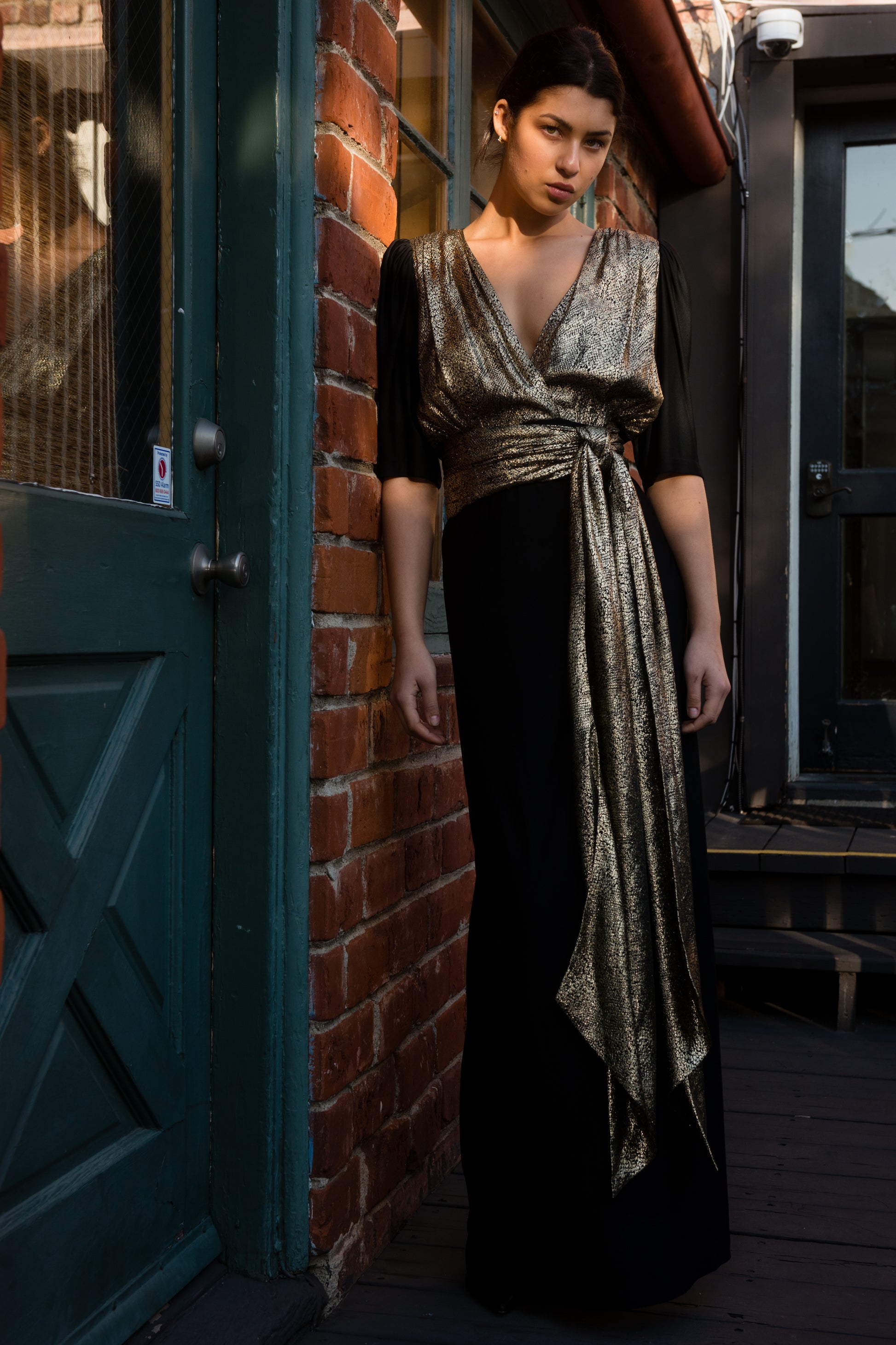 Woman in a black and gold dress standing against a brick wall.
