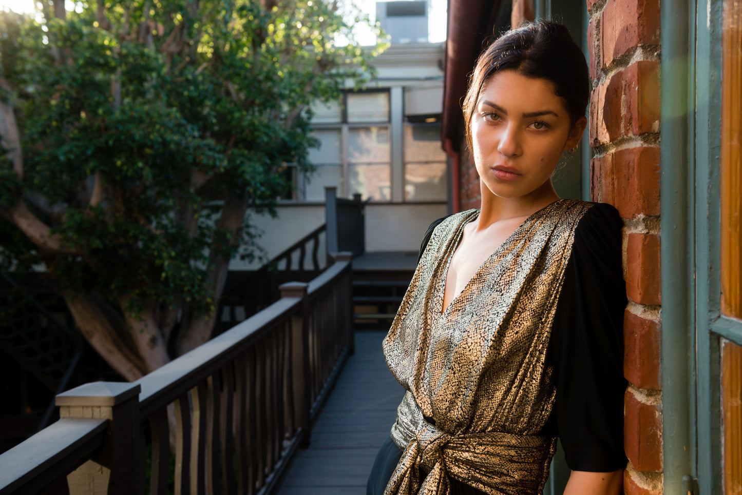 Woman standing on a balcony with a brick wall and greenery in the background