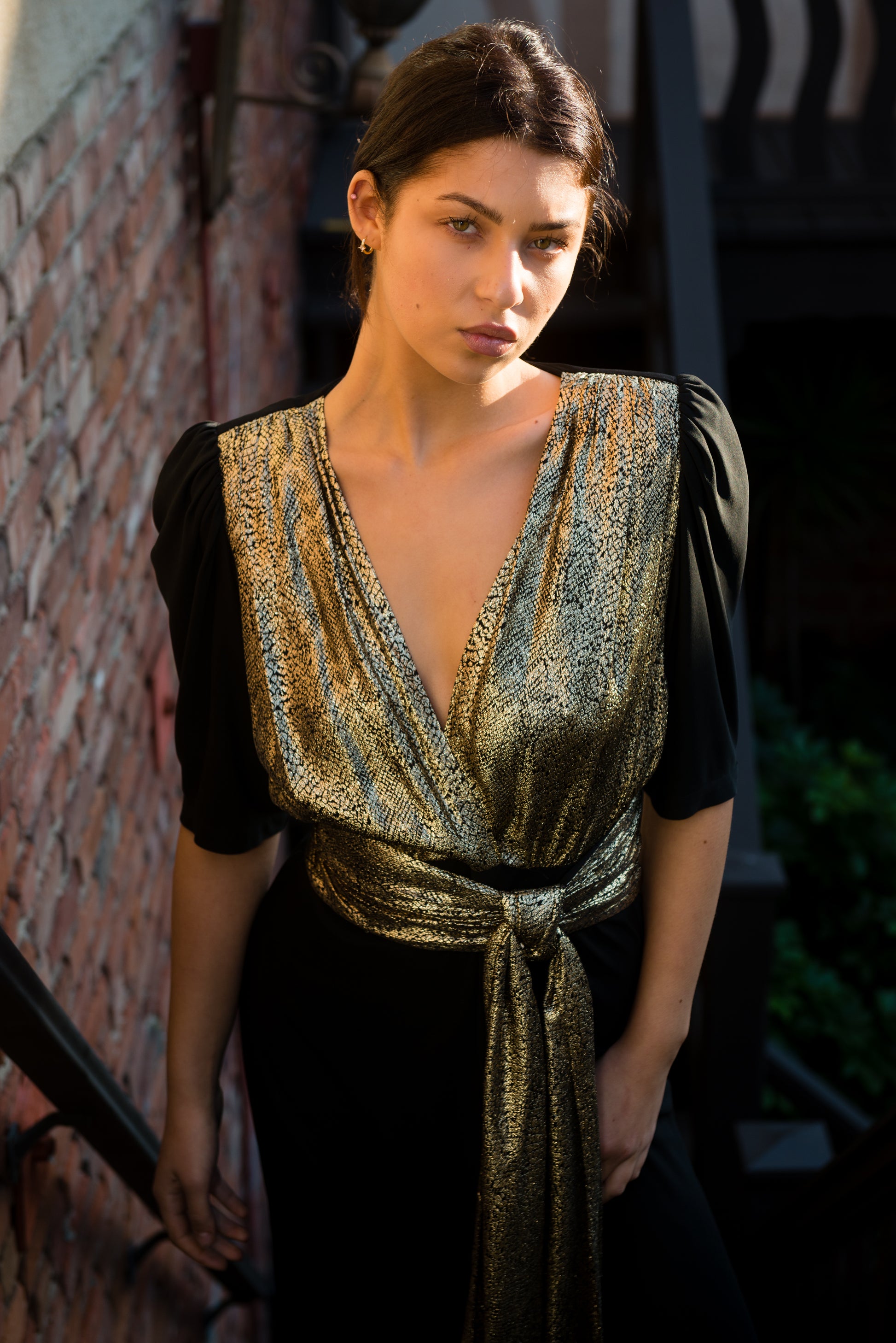 Woman wearing a gold and black dress against a brick wall.