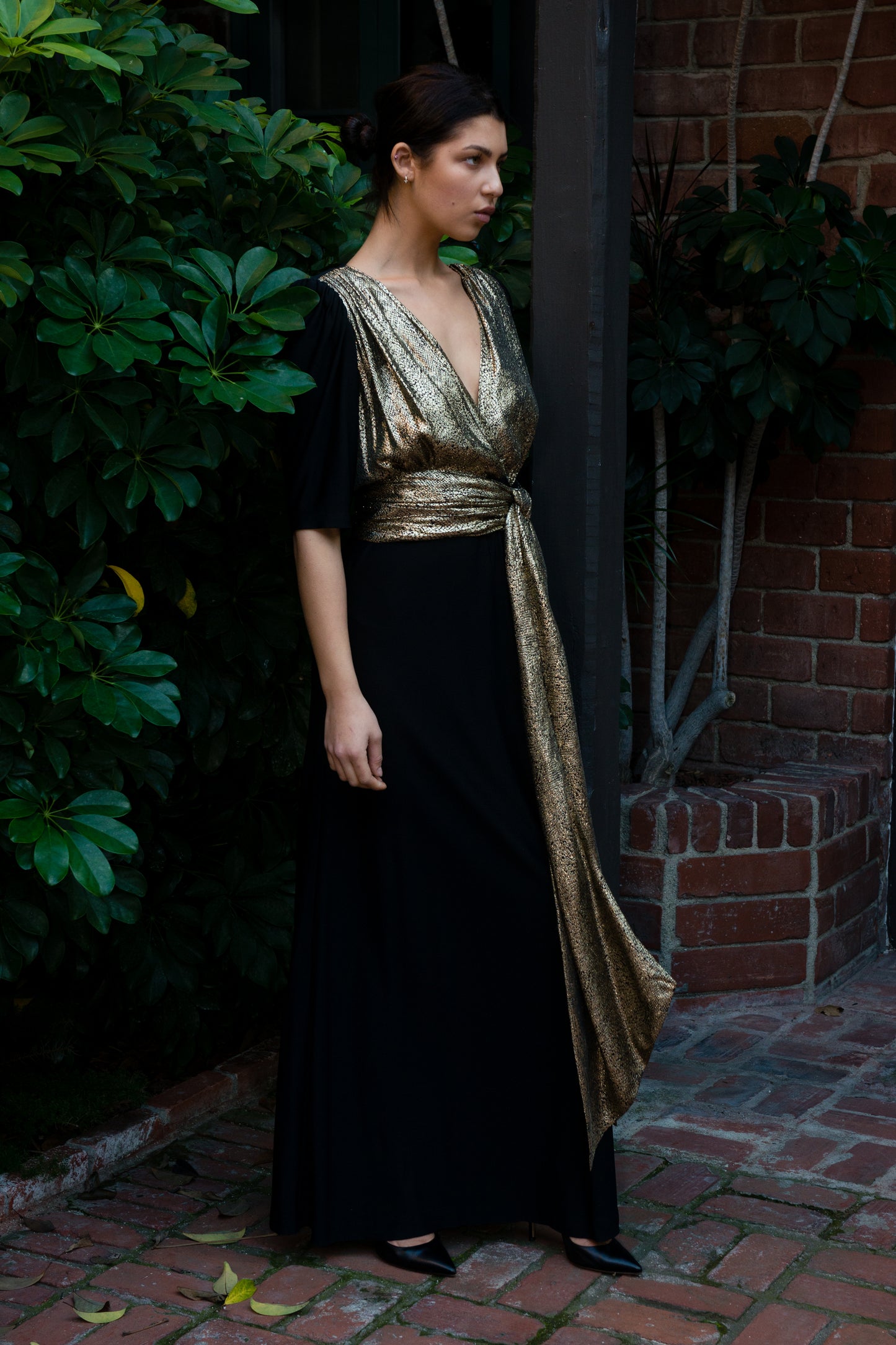 Woman wearing a black and gold dress standing against a brick wall with greenery.