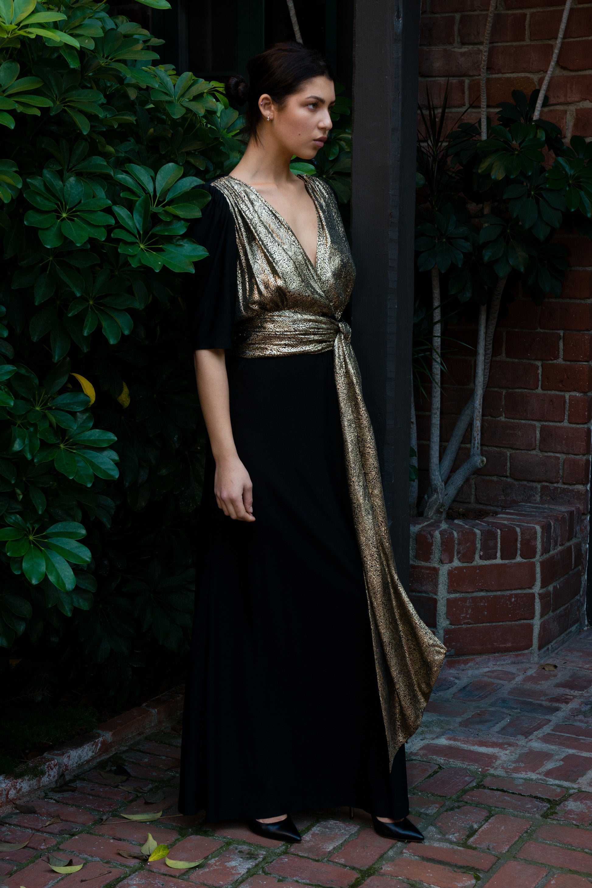 Woman wearing a black and gold dress standing against a brick wall with greenery.