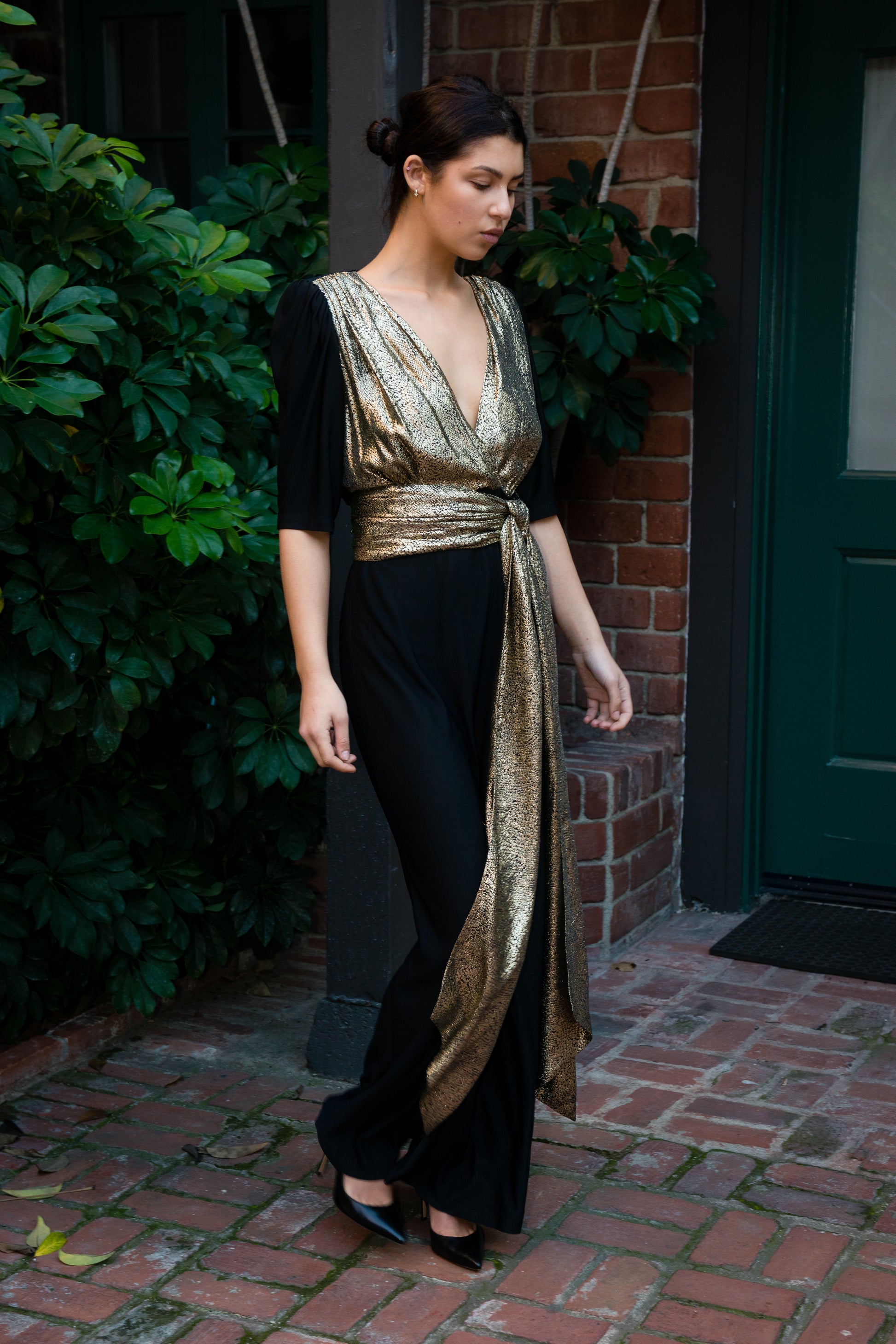 Woman wearing a black and gold outfit standing on a brick path with greenery and a door in the background.
