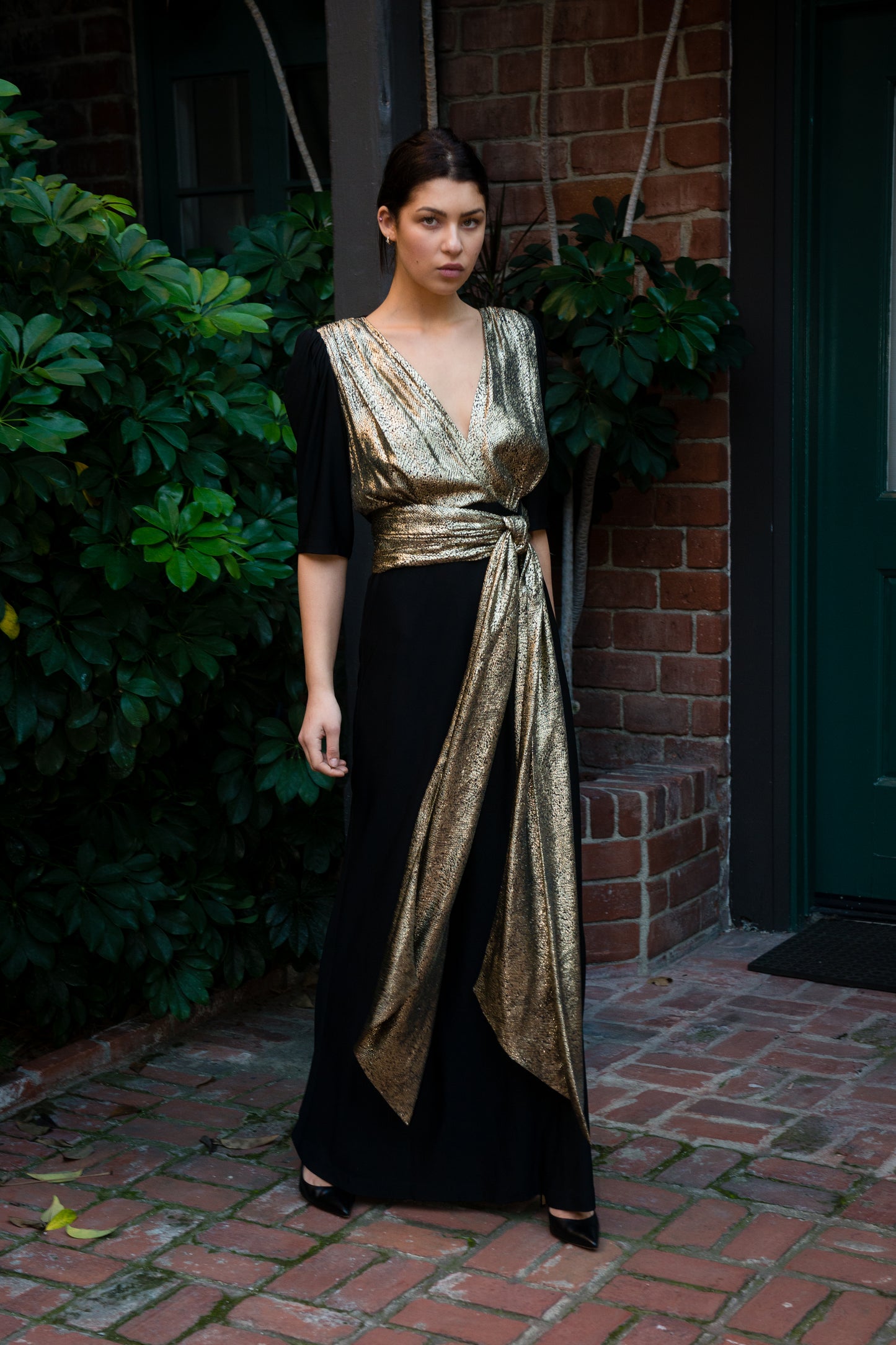 Woman wearing a black and gold dress standing outdoors against a brick wall.