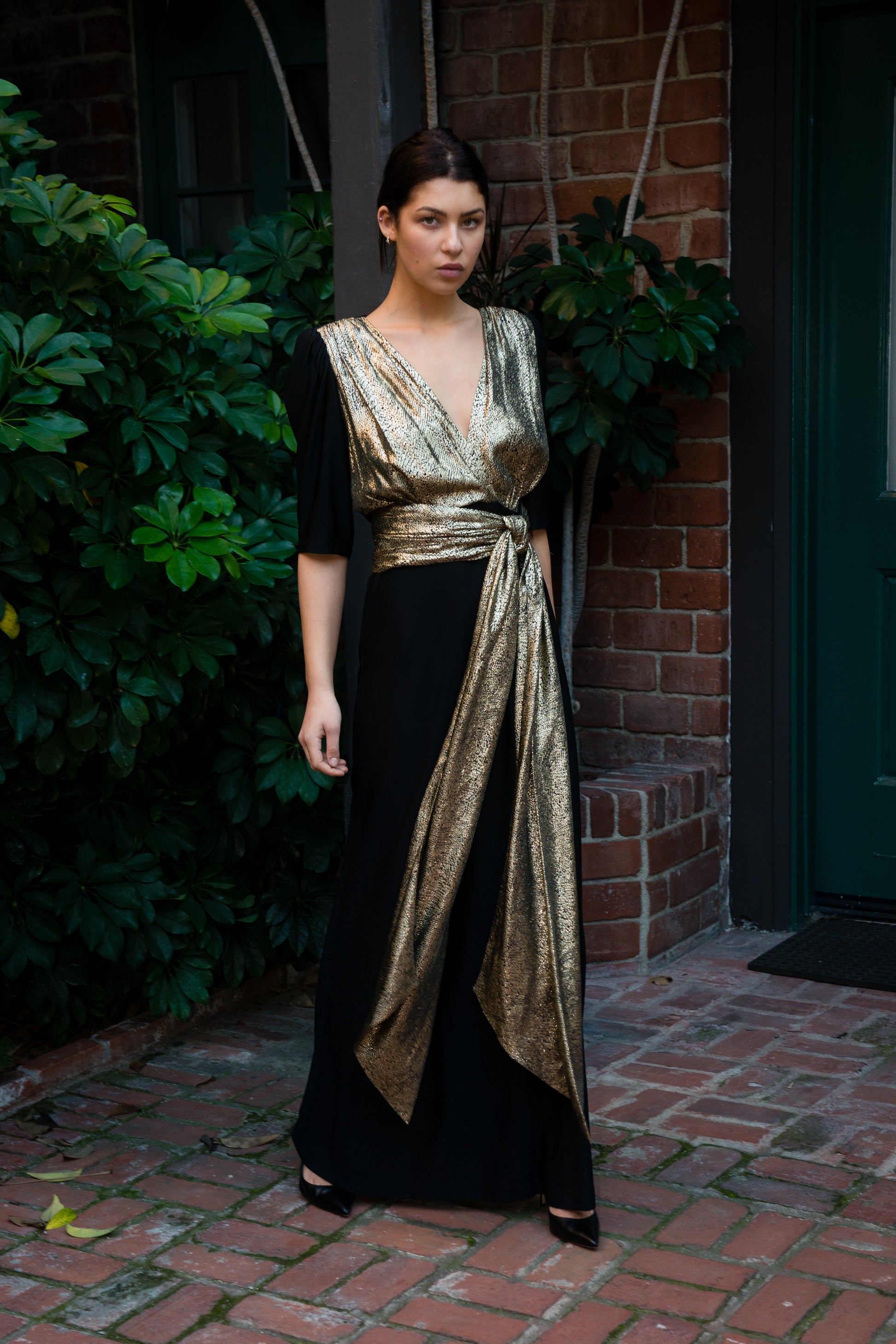 Woman wearing a black and gold dress standing outdoors against a brick wall.