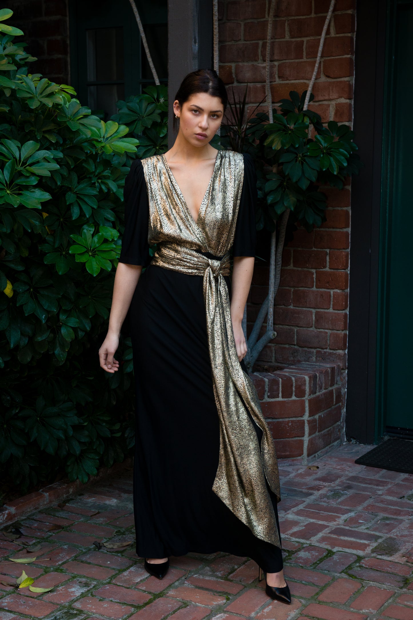 Woman wearing a black and gold dress standing outdoors against a brick wall.