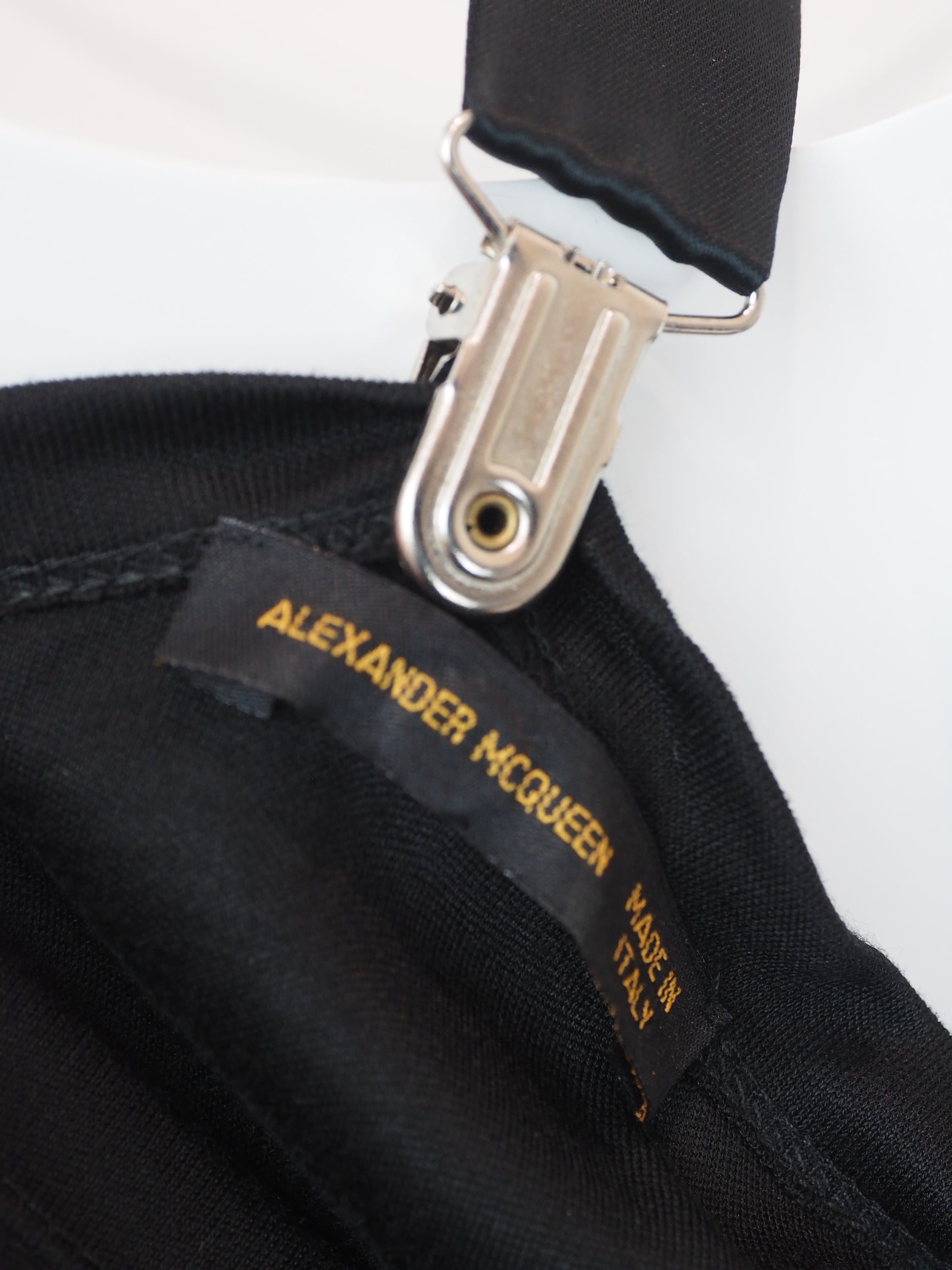 Close-up of a black fabric accessory with a black brand label reading Alexander McQueen in gold text, attached to a silver metal clip on a white background.