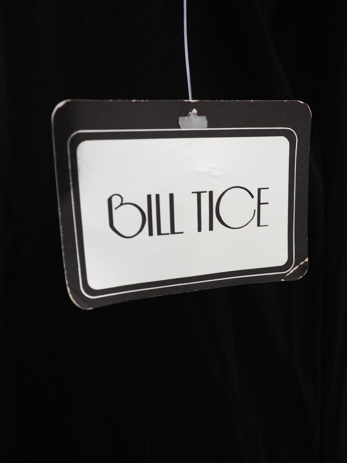 A hanging retail tag with the brand name 'BILL TICE' printed in black on a white label, attached to a black garment against a dark background.