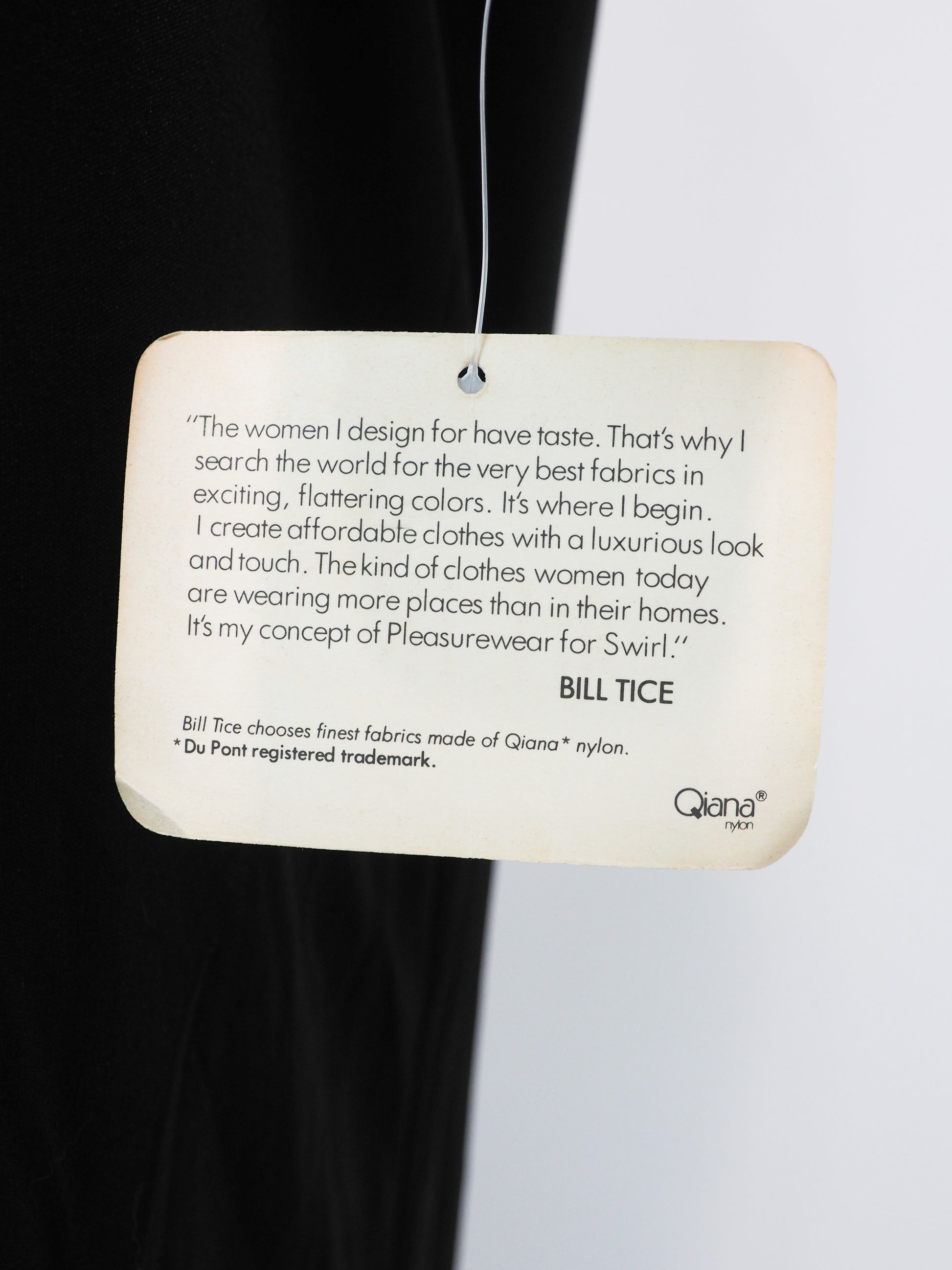 A cream-colored fabric care tag hanging from a thin wire against a split black and white background. The tag features printed text, including a quote by Bill Tice and the Qiana brand logo.