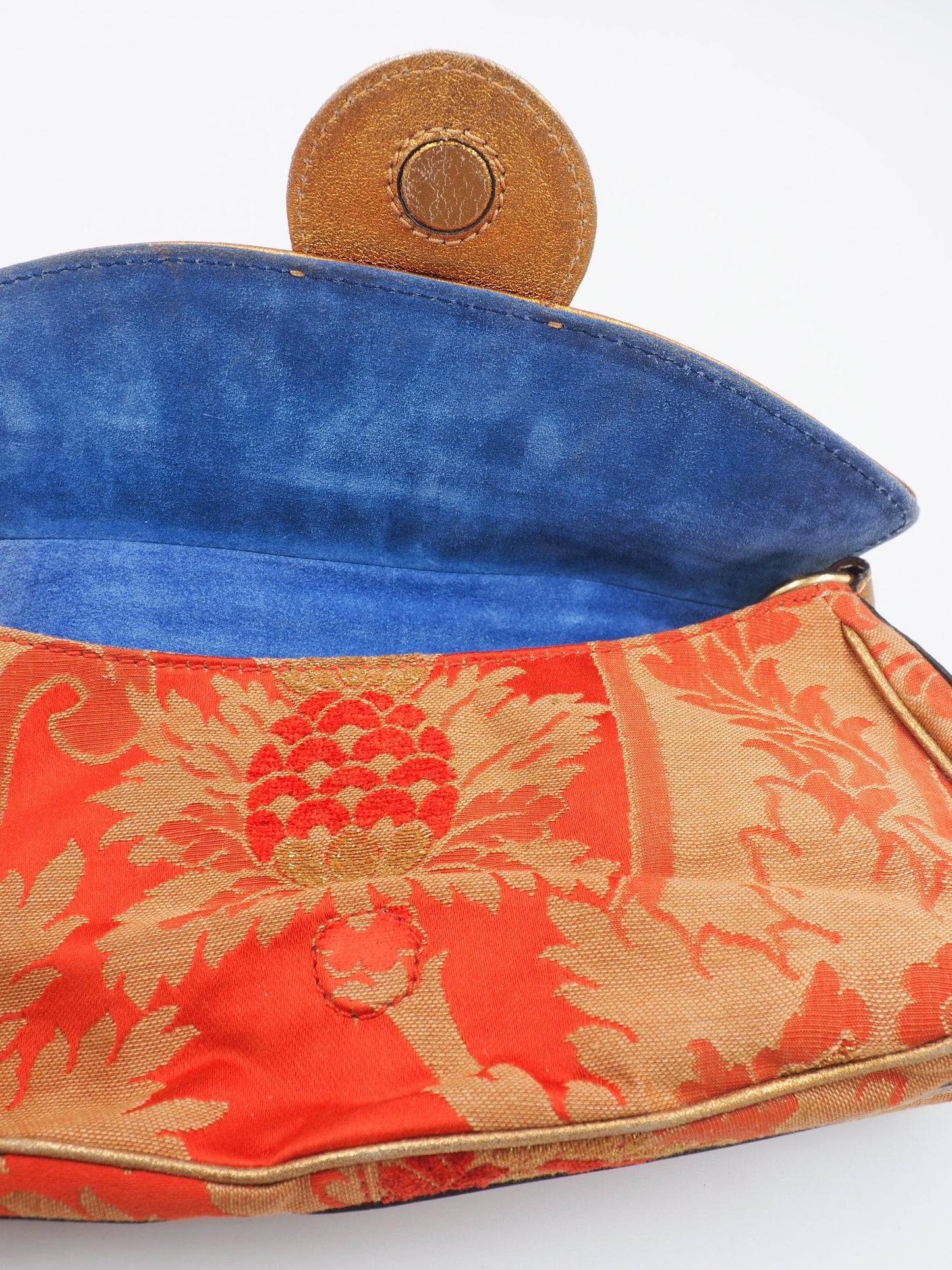 A close-up of a vintage etro handbag showing its interior, featuring a vibrant blue suede lining and a red-orange brocade fabric exterior with a gold pineapple motif and tan leather strap.
