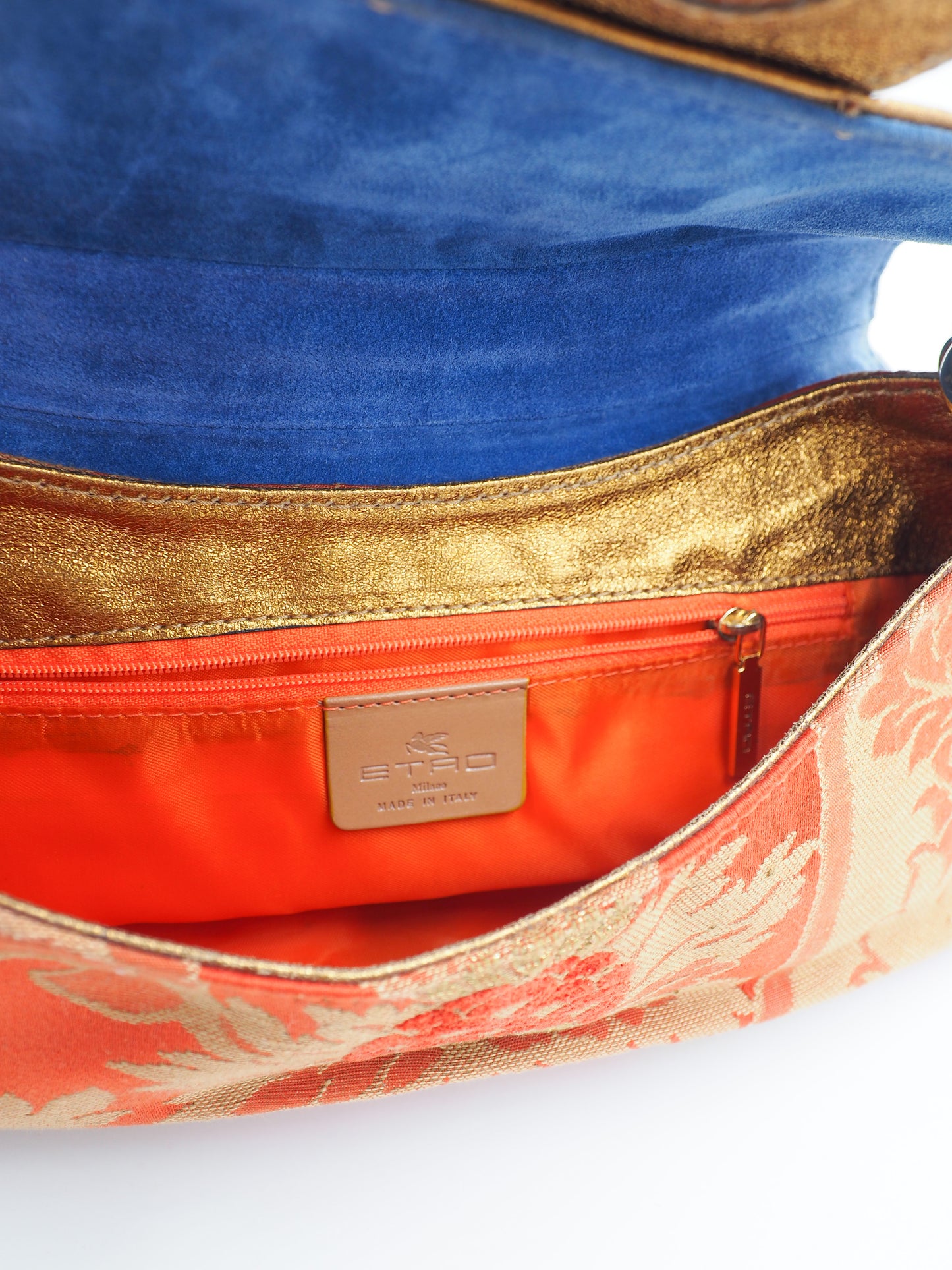 Close-up of a vintage etro handbag interior showing a blue velvet lining, a metallic gold trim, a bright orange zip pocket with a tan leather brand patch, and part of a peach and gold patterned exterior fabric.